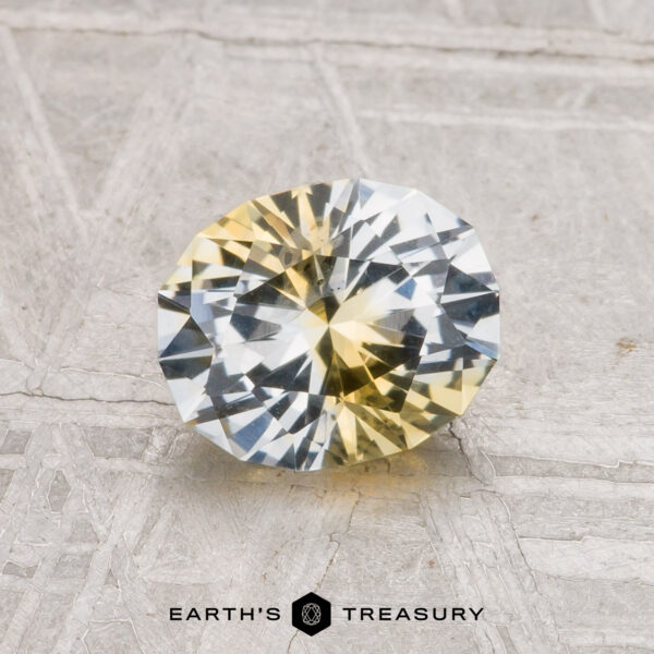 1.29-Carat Montana Sapphire (Heated) A 1.29-carat heated Montana sapphire, round and faceted with yellow and clear color zoning, rests on a textured gray surface. "Earth's Treasury" is shown at the bottom.