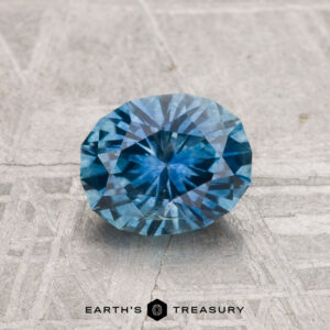 A 1.28-carat heated Montana sapphire with a round faceted cut is showcased on a textured gray surface, featuring the “EARTH’S TREASURY” text below.
