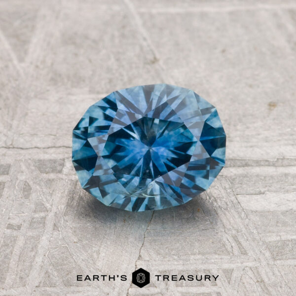 1.28-Carat Montana Sapphire (Heated) A 1.28-carat heated Montana sapphire with a round faceted cut is showcased on a textured gray surface, featuring the “EARTH’S TREASURY” text below.
