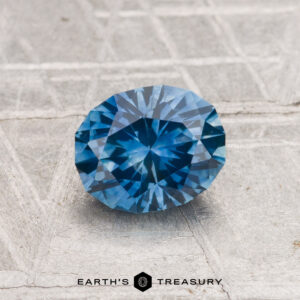 A 1.17-carat heated Montana sapphire with a brilliant cut is displayed on a textured gray surface. The text "EARTH'S TREASURY" appears at the bottom.