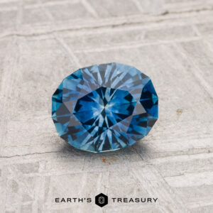 A 1.07-carat heated Montana sapphire, round and faceted, is displayed on a textured gray surface with the "EARTH'S TREASURY" logo at the bottom.