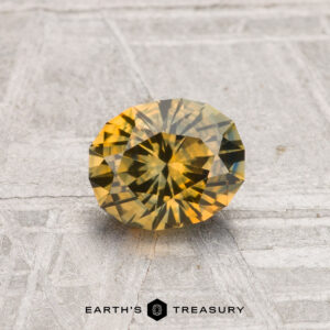 A round, faceted yellow-green 0.97-carat heated Montana sapphire is displayed on a textured gray surface with "EARTH'S TREASURY" below.