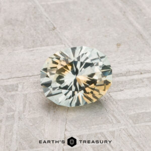 A 0.93-carat heated Montana sapphire with round, faceted cuts glimmers in light blue and yellow hues atop a textured gray surface bearing the "Earth's Treasury" logo.