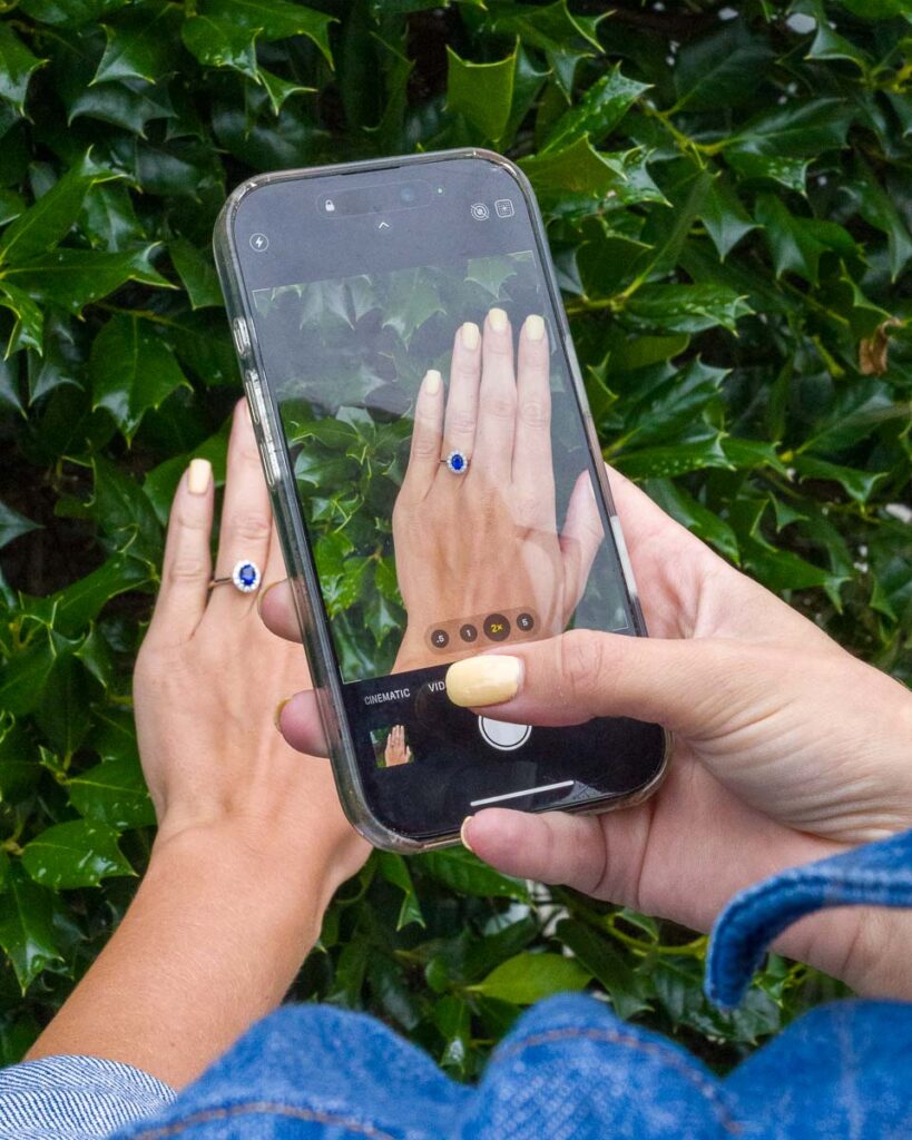 Someone photographs their hand wearing Taking Pictures of Your Ring 3, with green leaves in the background.