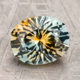 A faceted oval gem-color-particolored gemstone with yellow and blue-green hues rests on a textured gray surface, highlighting the vibrant beauty of all gemstones.