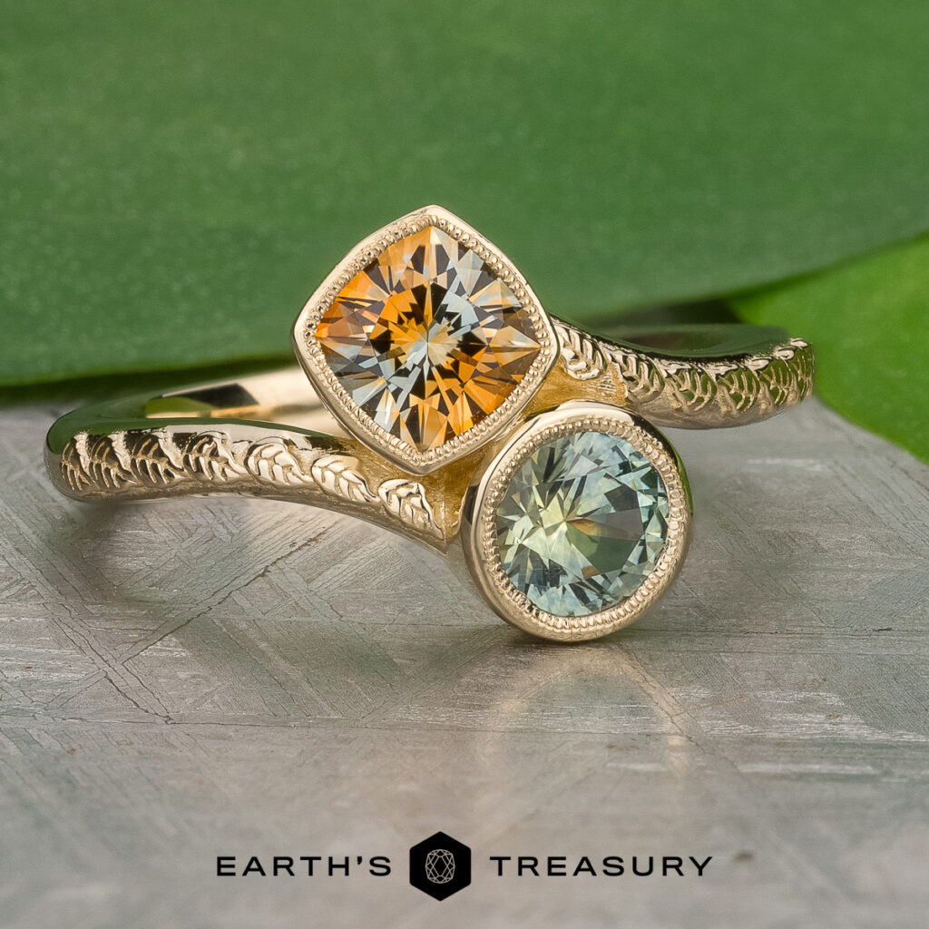 Two gold rings with engraved bands—one with a yellow gemstone, the other pale blue—are displayed side by side above the “Earth’s Treasury” logo. Product: Stacked from 15 images. Method=B (R=11,S=7).