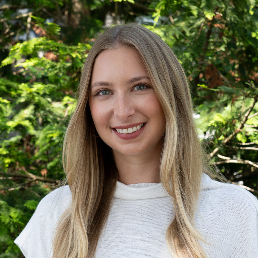 A smiling woman with long blonde hair, dressed in a white top, stands outdoors before green foliage. Photo: _DSC5935-Edit.