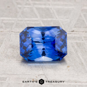 A 1.44-carat heated Ceylon sapphire with a rectangular, faceted cut is shown on a textured light gray surface, with the "EARTH'S TREASURY" logo and text beneath.
