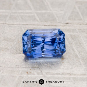 A 1.29-carat Ceylon sapphire (heated), radiant-cut and rectangular, sits on a textured gray surface with "EARTH'S TREASURY" visible below.