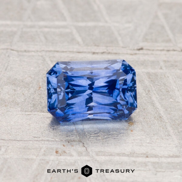 1.29-Carat Ceylon Sapphire (Heated) A 1.29-carat Ceylon sapphire (heated), radiant-cut and rectangular, sits on a textured gray surface with "EARTH'S TREASURY" visible below.
