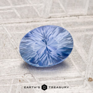 A 1.49-carat Ceylon sapphire with an oval cut and starburst facets rests on a textured metallic surface. "Earth's Treasury" appears at the bottom.