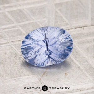 A faceted 1.45-carat Ceylon Sapphire with a starburst pattern rests on a textured gray surface, with the "Earth's Treasury" logo displayed at the bottom.
