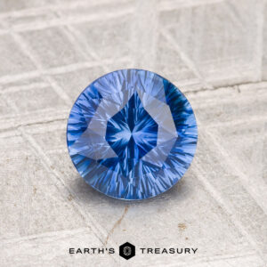 A 1.36-carat Ceylon Sapphire, round brilliant-cut and blue, is displayed on a textured light surface with "EARTH'S TREASURY" at the bottom.
