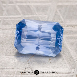 A 2.54-carat Ceylon sapphire with faceted edges sits on a textured gray surface; "EARTH'S TREASURY" appears at the bottom.