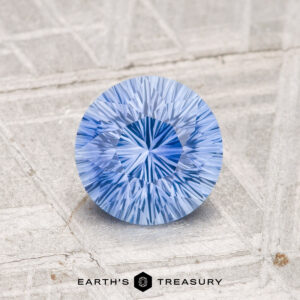 A 1.27-carat heated Ceylon sapphire with a round brilliant cut rests on a textured light gray surface, with the "EARTH'S TREASURY" logo and text below.
