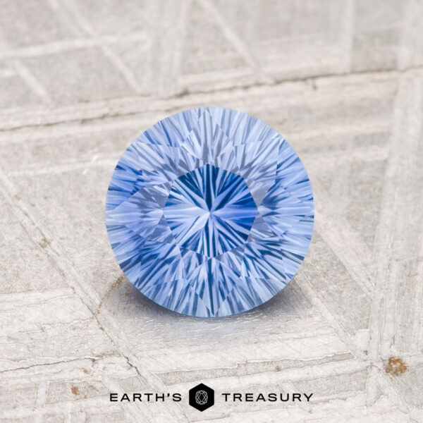 1.27-Carat Ceylon Sapphire (Heated) A 1.27-carat heated Ceylon sapphire with a round brilliant cut rests on a textured light gray surface, with the "EARTH'S TREASURY" logo and text below.