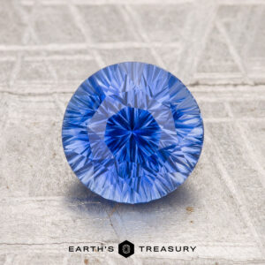 A 2.04-carat heated Ceylon sapphire, round and brilliant-cut, is showcased on a textured metallic surface with "Earth's Treasury" shown at the bottom.
