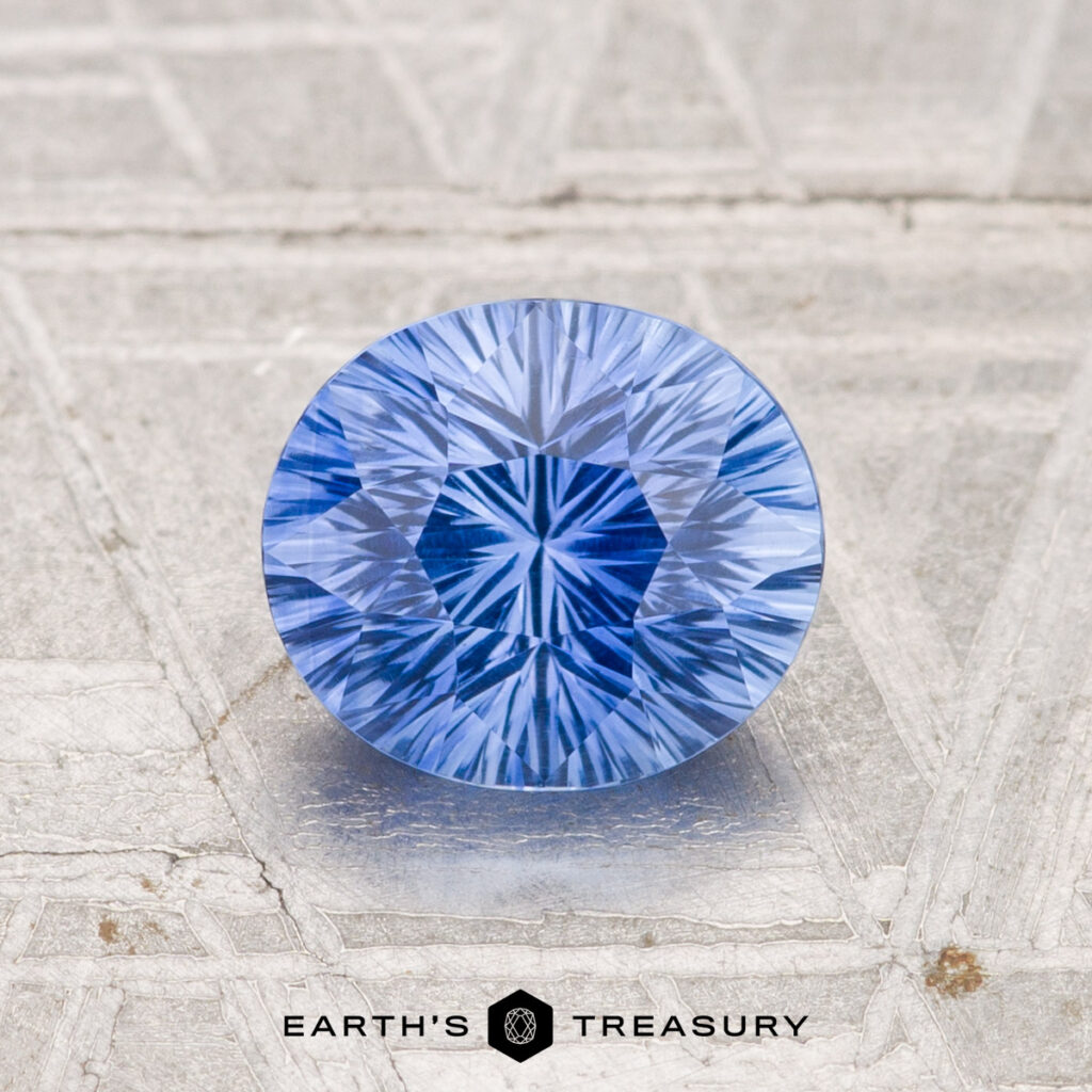 A round, faceted 1.45-Carat Ceylon Sapphire (Heated) in Cornflower Blue with a starburst pattern rests on a light, textured surface. The text "EARTH'S TREASURY" is shown at the bottom of the image.
