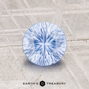 A 1.24-carat heated Ceylon sapphire, round and light blue with faceted cuts, sits on a textured light gray surface featuring the "EARTH'S TREASURY" logo at the bottom.