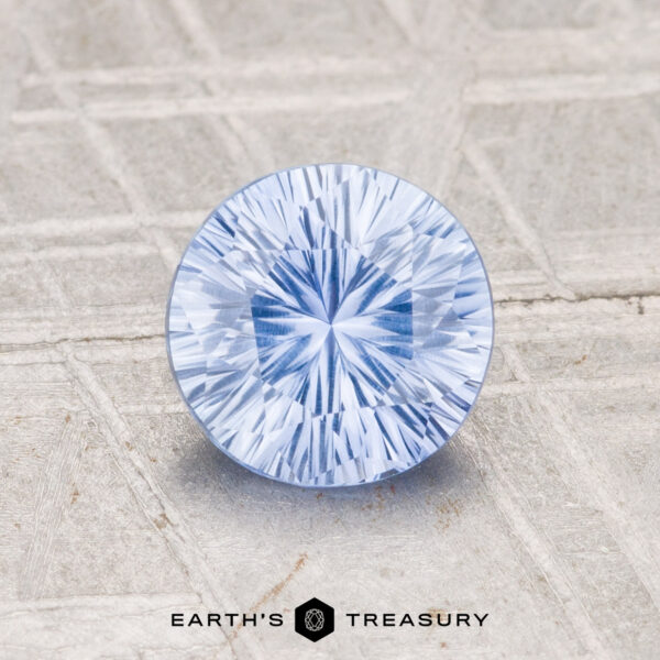 1.24-Carat Ceylon Sapphire (Heated) A 1.24-carat heated Ceylon sapphire, round and light blue with faceted cuts, sits on a textured light gray surface featuring the "EARTH'S TREASURY" logo at the bottom.
