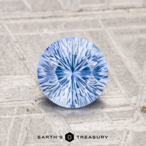 A 1.05-carat, brilliantly-cut Ceylon sapphire (heated) rests on a textured gray surface with the "EARTH'S TREASURY" logo at the bottom.