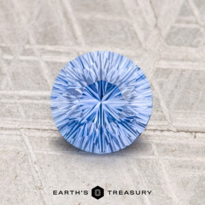A round, brilliant-cut 1.26-Carat Ceylon Sapphire (Heated) rests on a textured light gray surface, with "EARTH’S TREASURY" displayed below.