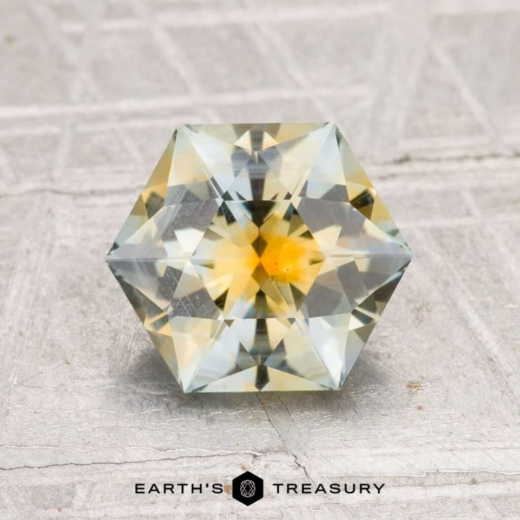 A 2.09-carat faceted hexagonal Montana Sapphire with yellow and green hues is displayed upright on a textured gray surface with "EARTH'S TREASURY" text below.