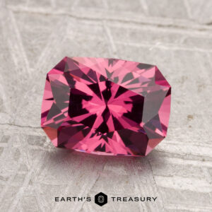 A 2.24-carat faceted rectangular Tanga Garnet gemstone in vivid pink is displayed on a textured light gray surface with "EARTH'S TREASURY" text at the bottom.
