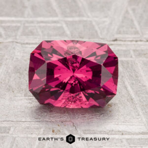 A 2.22-carat Tanga Garnet, rectangular and faceted in deep pink, rests on a textured, light-colored surface with the "Earth's Treasury" logo beneath.