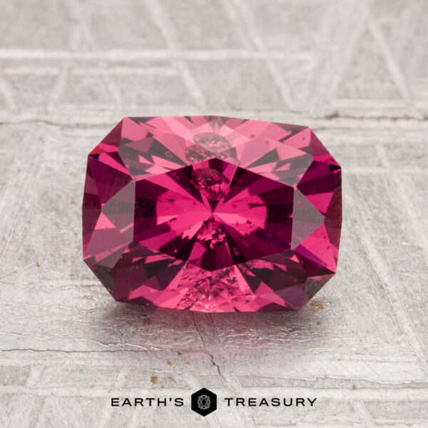 2.22-Carat Tanga Garnet A 2.22-carat Tanga Garnet, rectangular and faceted in deep pink, rests on a textured, light-colored surface with the "Earth's Treasury" logo beneath.