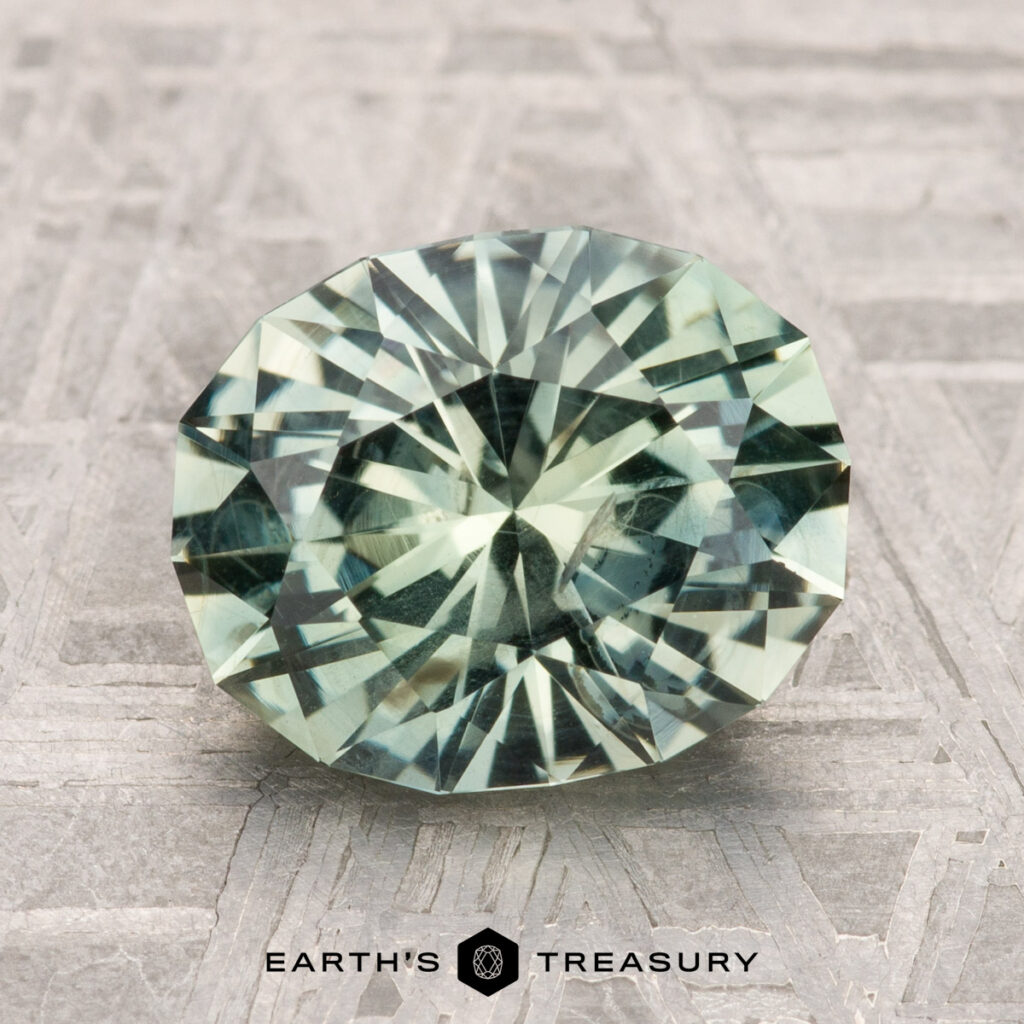 A 3.25-carat Montana Sapphire, faceted in an oval shape with a green hue, is displayed on a patterned gray surface with the text "Earth's Treasury" below.
