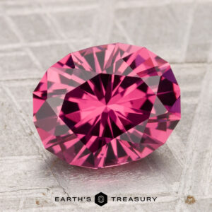 A 3.34-carat Tanga Garnet, oval-cut and deep pink with intricate facets, is displayed on a textured gray surface with the text "EARTH'S TREASURY" at the bottom.