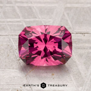 A faceted, rectangular 2.03-carat Tanga Garnet gemstone is displayed on a textured gray surface, with "EARTH'S TREASURY" at the bottom.