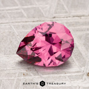 A 1.95-carat faceted, pear-shaped Tanga Garnet gemstone rests on a textured gray surface, with the "EARTH'S TREASURY" logo at the bottom of the image.