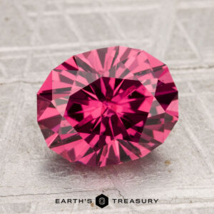 A 3.27-carat Tanga Garnet, round with a brilliant faceted cut, is displayed on a textured gray surface with the "Earth's Treasury" logo at the bottom.