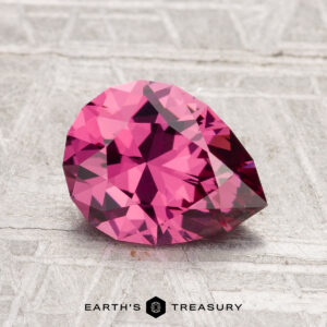 A 1.73-carat Tanga Garnet, pear-shaped and faceted in pink, sits on a textured gray surface with "EARTH'S TREASURY" displayed at the bottom.