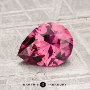 A faceted, pear-shaped 1.54-carat Tanga Garnet gemstone rests on a textured, light-colored surface with "Earth's Treasury" printed below.