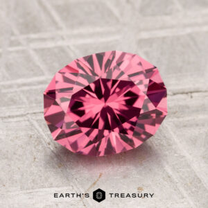 A 2.36-carat Tanga Garnet, round and brilliant-cut, is showcased on a textured gray surface with "EARTH'S TREASURY" displayed below.