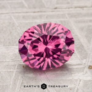A 2.33-carat Tanga Garnet, faceted in an oval shape and displaying a vibrant pink hue, rests on a textured light surface with the "Earth's Treasury" logo and text displayed below.