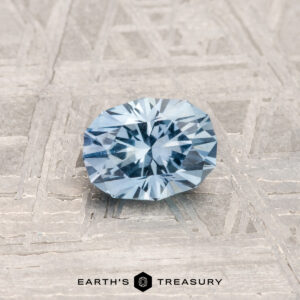 A 0.72-carat, round brilliant Montana sapphire sits on a textured metal surface with "EARTH'S TREASURY" displayed at the bottom.