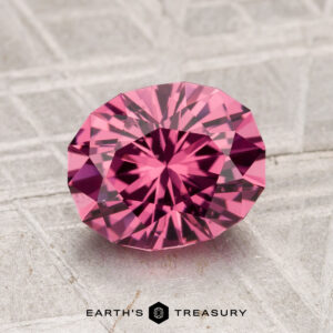 A 2.21-carat Tanga Garnet, faceted in an oval shape and displaying a vivid pink hue, is showcased on a textured gray surface with the "Earth's Treasury" logo at the bottom.