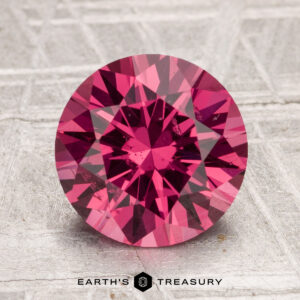 A round, brilliant-cut 3.04-carat Tanga garnet in deep pink is displayed upright on a textured gray surface with "Earth's Treasury" text below.