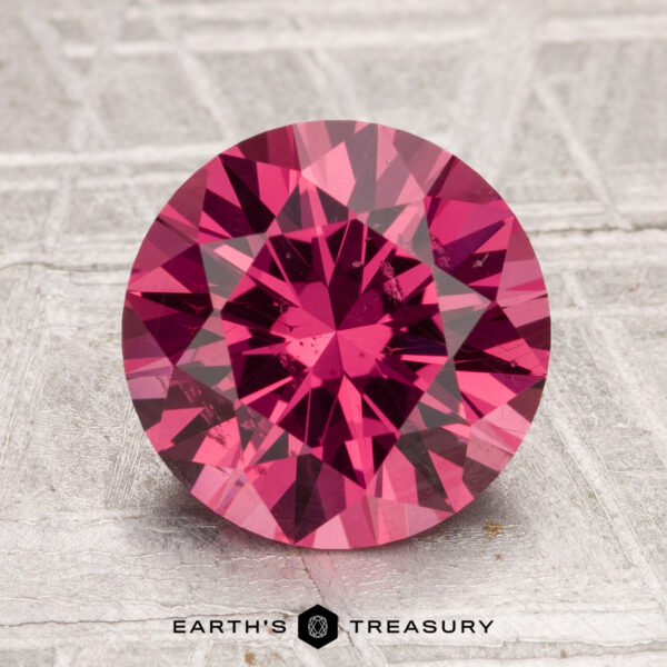A round, brilliant-cut 3.04-carat Tanga garnet in deep pink is displayed upright on a textured gray surface with "Earth's Treasury" text below.