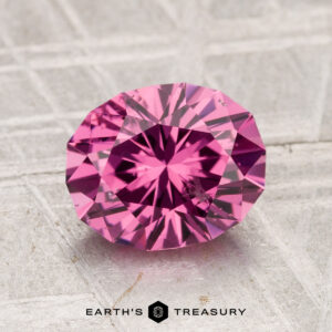 A 2.18-carat Tanga Garnet, cut in a faceted oval shape and displaying a vivid pink hue, rests on a textured light gray surface with the text "EARTH'S TREASURY" below it.