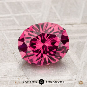 A 2.01-carat Tanga Garnet, cut in a faceted oval shape and displaying a vivid pink hue, rests on a textured gray surface with "EARTH'S TREASURY" shown at the bottom.