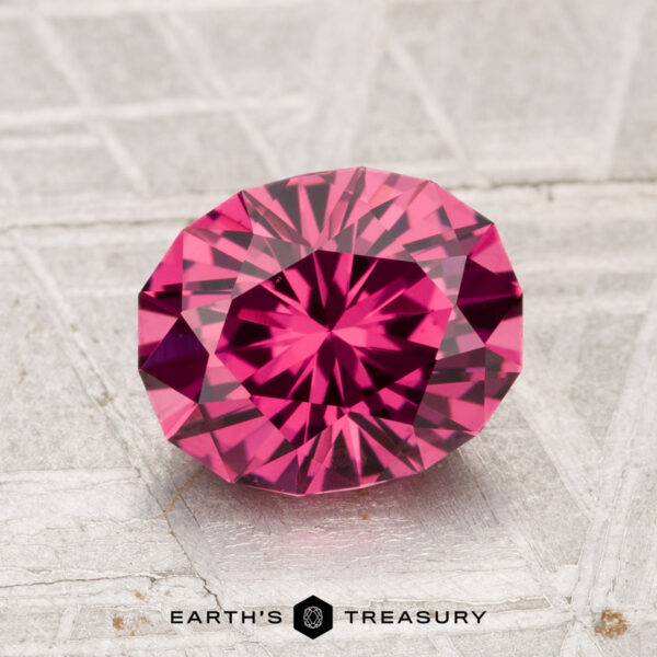 2.01-Carat Tanga Garnet A 2.01-carat Tanga Garnet, cut in a faceted oval shape and displaying a vivid pink hue, rests on a textured gray surface with "EARTH'S TREASURY" shown at the bottom.