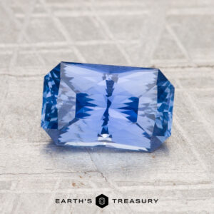 A 2.07-carat rectangular step-cut Ceylon sapphire is shown on a textured gray surface, with "EARTH'S TREASURY" displayed at the bottom.