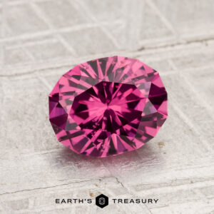A 1.74-carat faceted, oval-cut Tanga Garnet gemstone rests on a textured gray surface, with the text "EARTH'S TREASURY" visible below it.