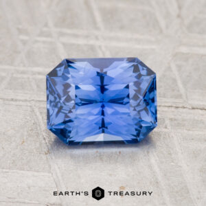 A 1.41-carat, rectangular cornflower blue Ceylon Sapphire rests on a light textured surface with "EARTH'S TREASURY" below.