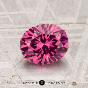 A 1.65-carat Tanga Garnet, round brilliant-cut with sharp facets, is displayed on a textured gray surface with the Earth’s Treasury logo beneath it.